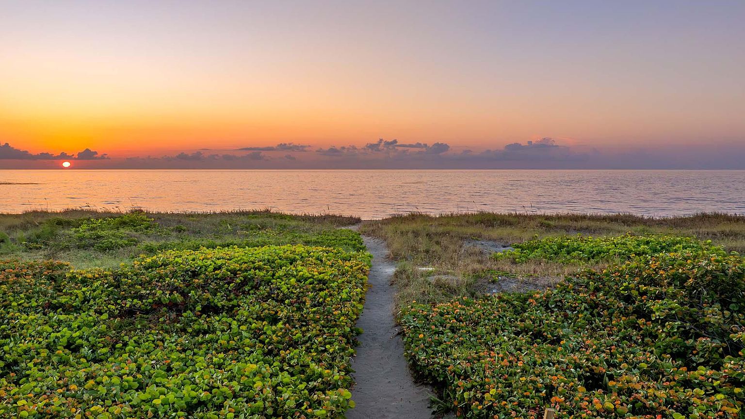 This image showcases a serene beach scene at sunset. A sandy path leads from lush green vegetation towards the ocean, where the sun is setting, casting a warm orange glow across the water and sky. The scene evokes a sense of tranquility and natural beauty, highlighting the property's proximity to the beach.