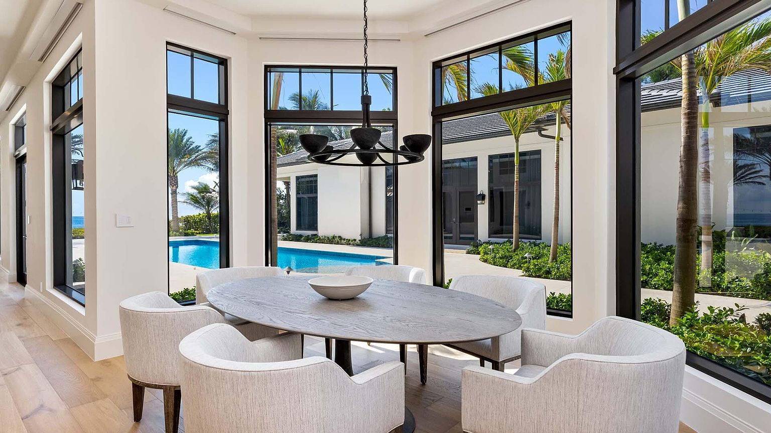This is an interior shot of a dining room featuring an oval wooden table surrounded by six upholstered chairs. Large windows with black frames offer views of a pool, palm trees, and a modern white building, creating a bright and airy atmosphere. A modern chandelier hangs above the table, adding a touch of elegance.