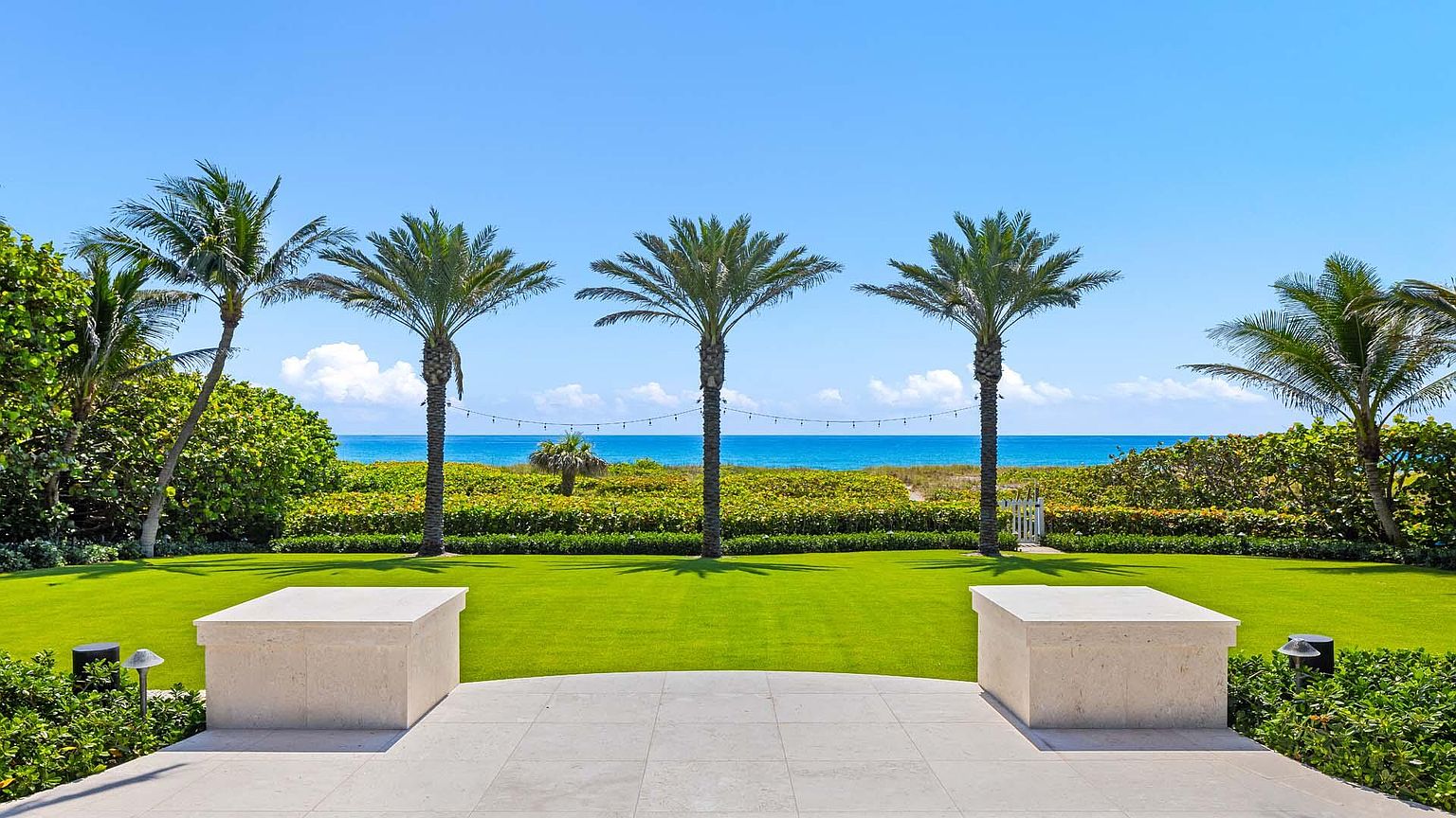This image showcases a meticulously maintained backyard with a lush green lawn, framed by palm trees and manicured hedges. The view extends to a serene ocean backdrop under a clear blue sky, creating a tranquil and luxurious atmosphere. The patio area in the foreground features elegant stone elements, enhancing the overall appeal of the outdoor space.