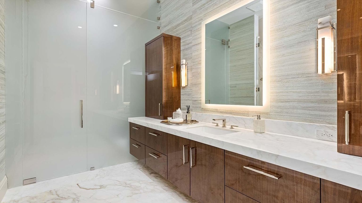 This is a primary bathroom featuring a modern design with a glass-enclosed shower and a long vanity with dark wood cabinets and a white countertop. The illuminated mirror and sconces provide ample lighting, while the marble flooring adds a touch of luxury. The perspective is a wide shot, showcasing the entire vanity area and the shower.