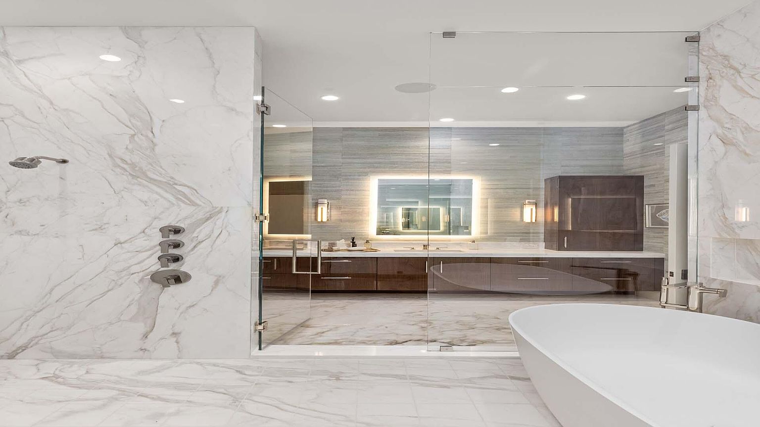 This is a luxurious primary bathroom featuring marble walls and flooring, a glass-enclosed shower with modern fixtures, and a freestanding white bathtub. The vanity area has dark wood cabinetry, illuminated mirrors, and stylish lighting. The overall impression is one of elegance and spa-like tranquility.
