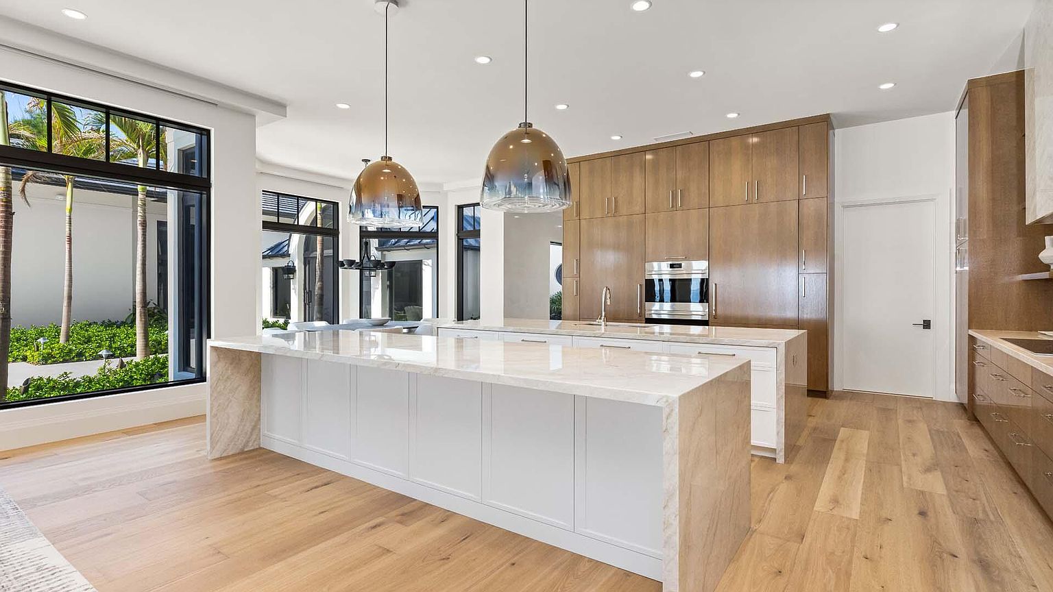 This is a bright and modern kitchen featuring a large island with a white base and marble countertop. The kitchen includes wood cabinets, stainless steel appliances, and hardwood floors. Large windows provide natural light and a view of the exterior landscaping.