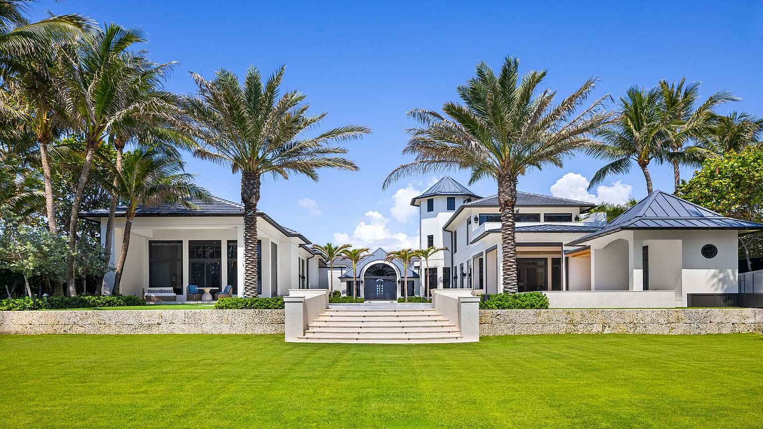 This is a front view of a luxurious, modern beachfront property. The house features a symmetrical design with a central tower, flanked by wings with dark-framed windows and dark-tiled roofs. Palm trees line the property, and a manicured lawn leads up to a stone retaining wall and a grand staircase to the entrance.