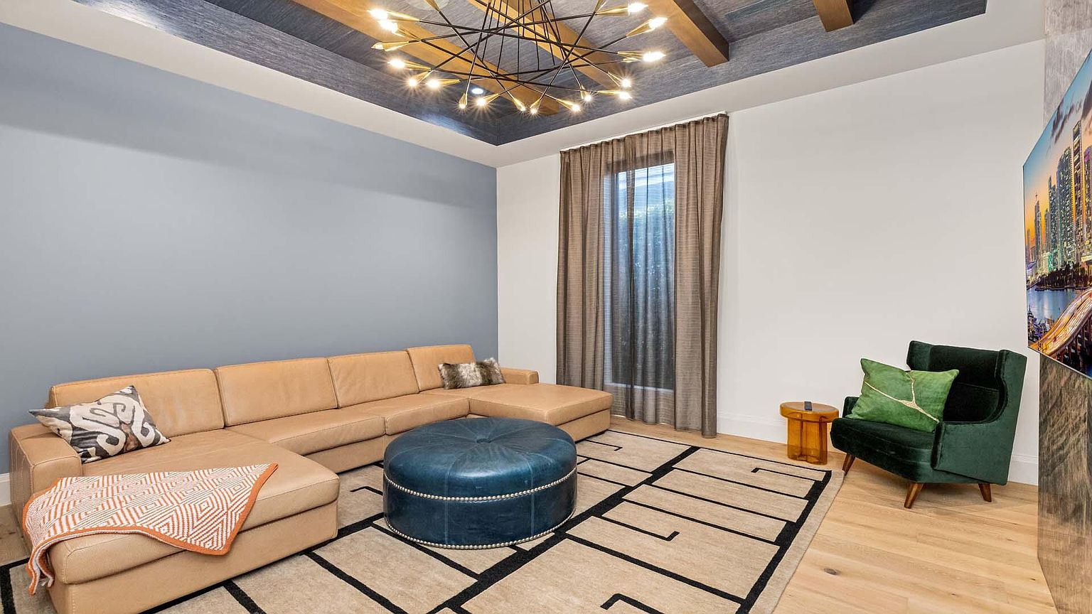 This is an interior shot of a living room featuring a large, tan sectional sofa, a blue leather ottoman, and a patterned area rug. A modern chandelier hangs from the textured ceiling, and natural light filters through the window with brown curtains. The room is decorated with throw pillows and a small side table, creating a cozy and inviting atmosphere.