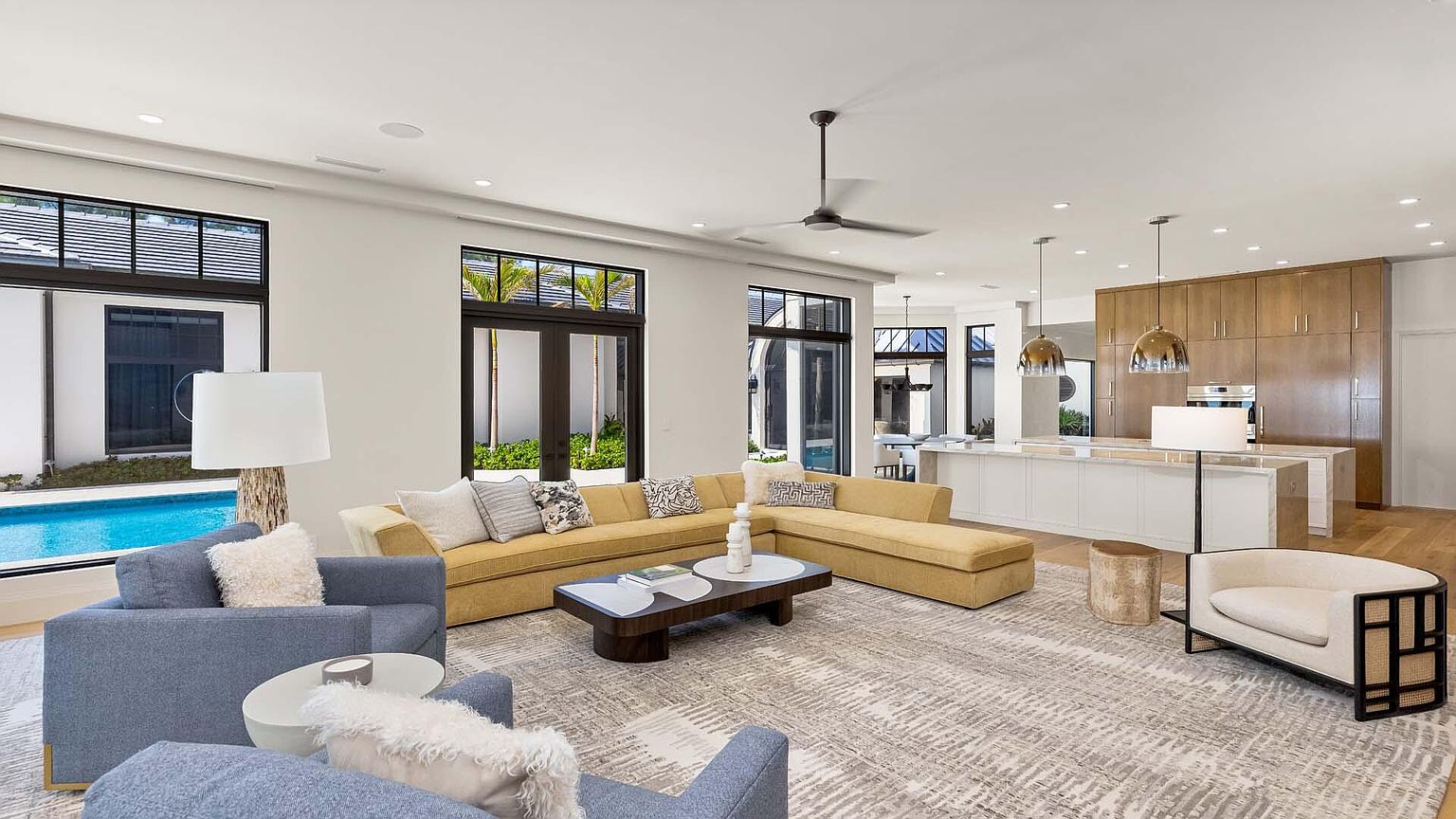 This is a spacious and modern living room with an open floor plan that seamlessly connects to the kitchen. The room features a large sectional sofa, comfortable armchairs, and a stylish coffee table, all arranged on a patterned area rug. Large windows provide ample natural light and views of the outdoor pool area, creating a bright and inviting atmosphere.