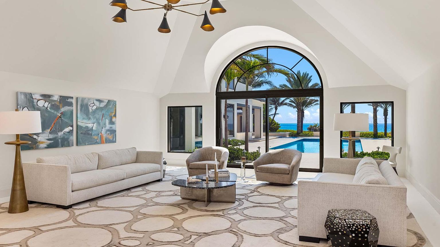 This is a bright and airy living room with high vaulted ceilings and a large arched window offering a view of a pool and the ocean. The room features neutral-toned sofas, swivel chairs, and a patterned rug, creating a sophisticated and comfortable atmosphere. Abstract artwork adorns the walls, and a modern chandelier hangs from the ceiling, adding to the room's elegant design.