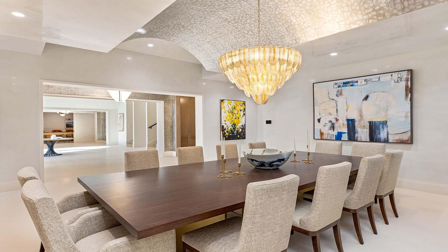This is an interior shot of a dining room featuring a large wooden table surrounded by upholstered chairs. A grand chandelier hangs above the table, and artwork adorns the walls. The room has a sophisticated and elegant feel, with a neutral color palette and ample natural light, creating an inviting space for dining and entertaining.