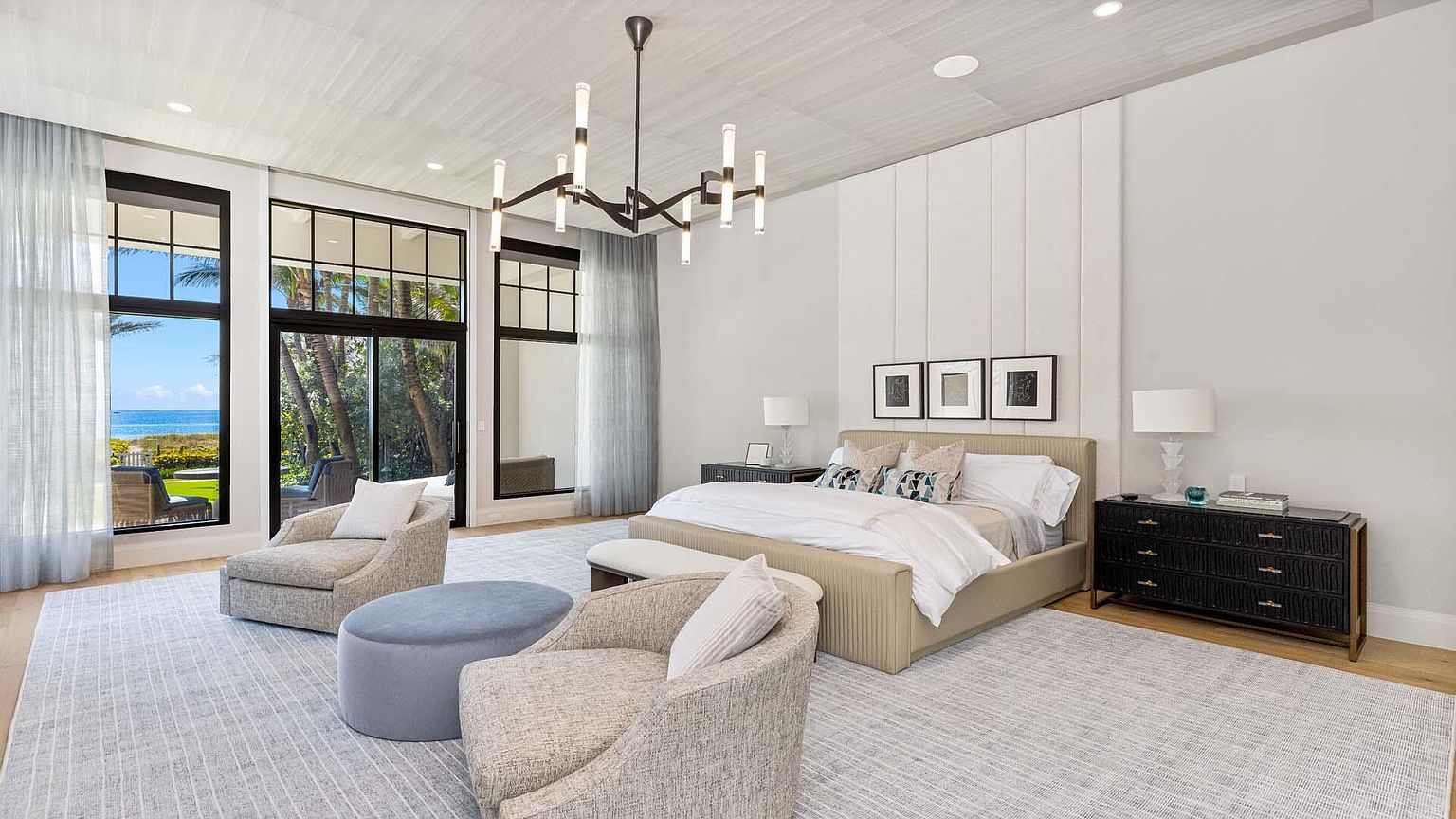 This is a luxurious primary bedroom featuring a neutral color palette, a large bed with decorative pillows, and a modern chandelier. The room has large windows offering a view of the ocean, and comfortable seating is arranged near the windows. The overall impression is one of elegance and tranquility.