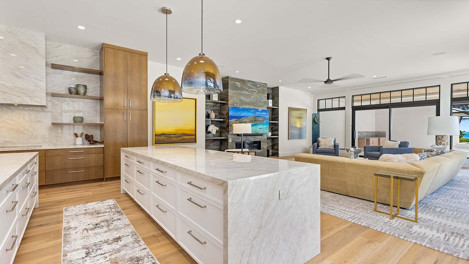 This is a bright and open kitchen and living area featuring a large marble island with white cabinetry and modern hardware. The kitchen flows seamlessly into the living room, which has a comfortable beige sectional sofa and large sliding glass doors. The space is well-lit with natural light and recessed lighting, creating a welcoming and luxurious atmosphere.
