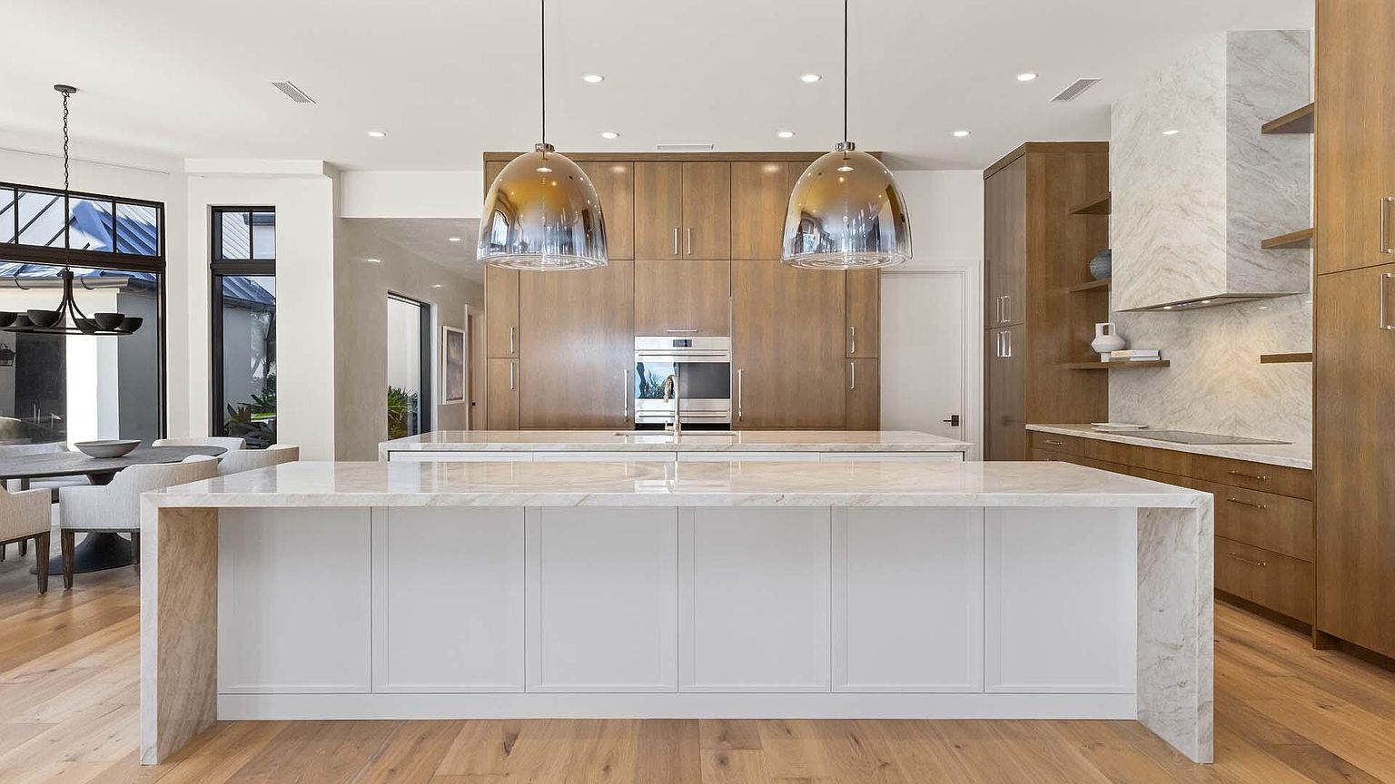 This is a bright and modern kitchen featuring a large island with a white base and marble countertop, complemented by wooden cabinetry and stainless steel appliances. Two pendant lights hang above the island, adding a touch of elegance. The kitchen has an open layout, with a dining area visible in the background, creating a seamless flow between spaces.