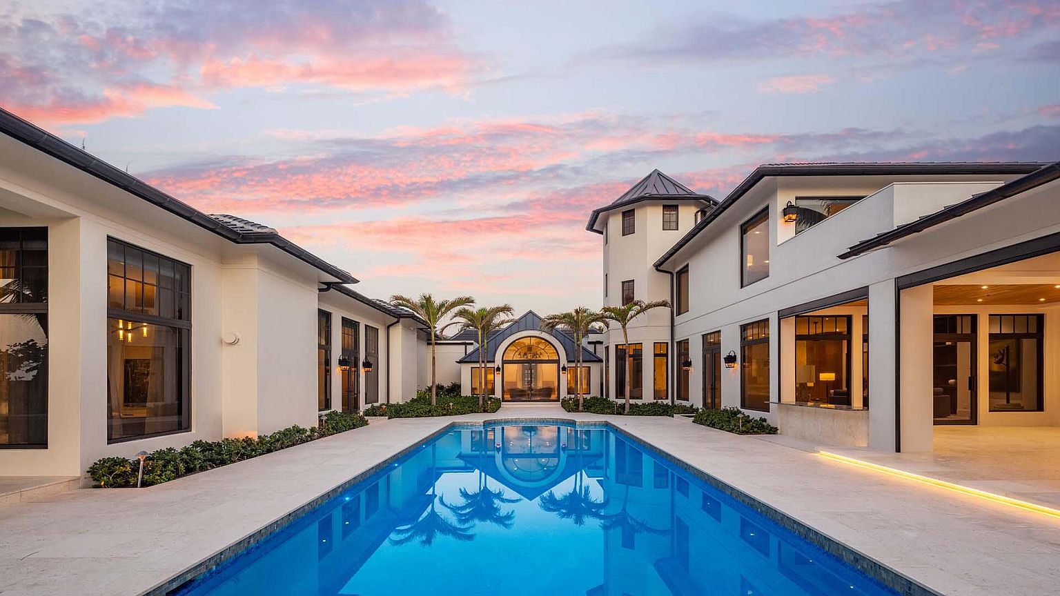 This image showcases a luxurious outdoor pool area of a modern estate. The rectangular pool reflects the architecture of the surrounding buildings and the colorful sky at dusk. Palm trees line the walkway, adding to the tropical ambiance, while the clean lines and elegant design of the buildings suggest high-end living.
