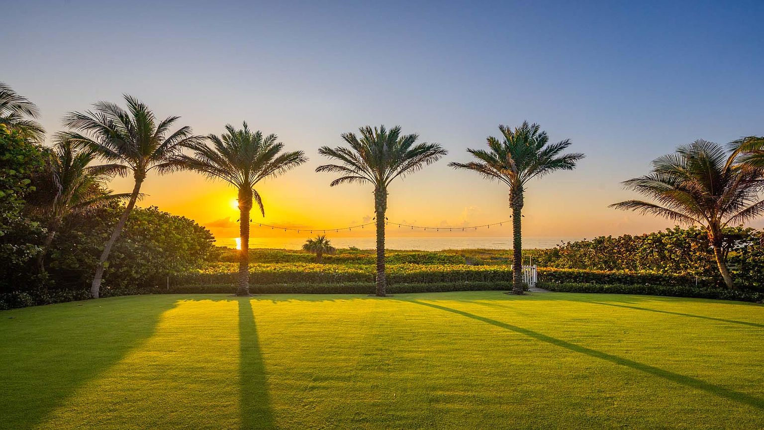 This exterior shot showcases a lush, well-manicured lawn leading to a beachfront property at sunset. Palm trees line the horizon, creating a tropical and serene atmosphere. The scene evokes a sense of luxury and relaxation, perfect for a high-end real estate listing.