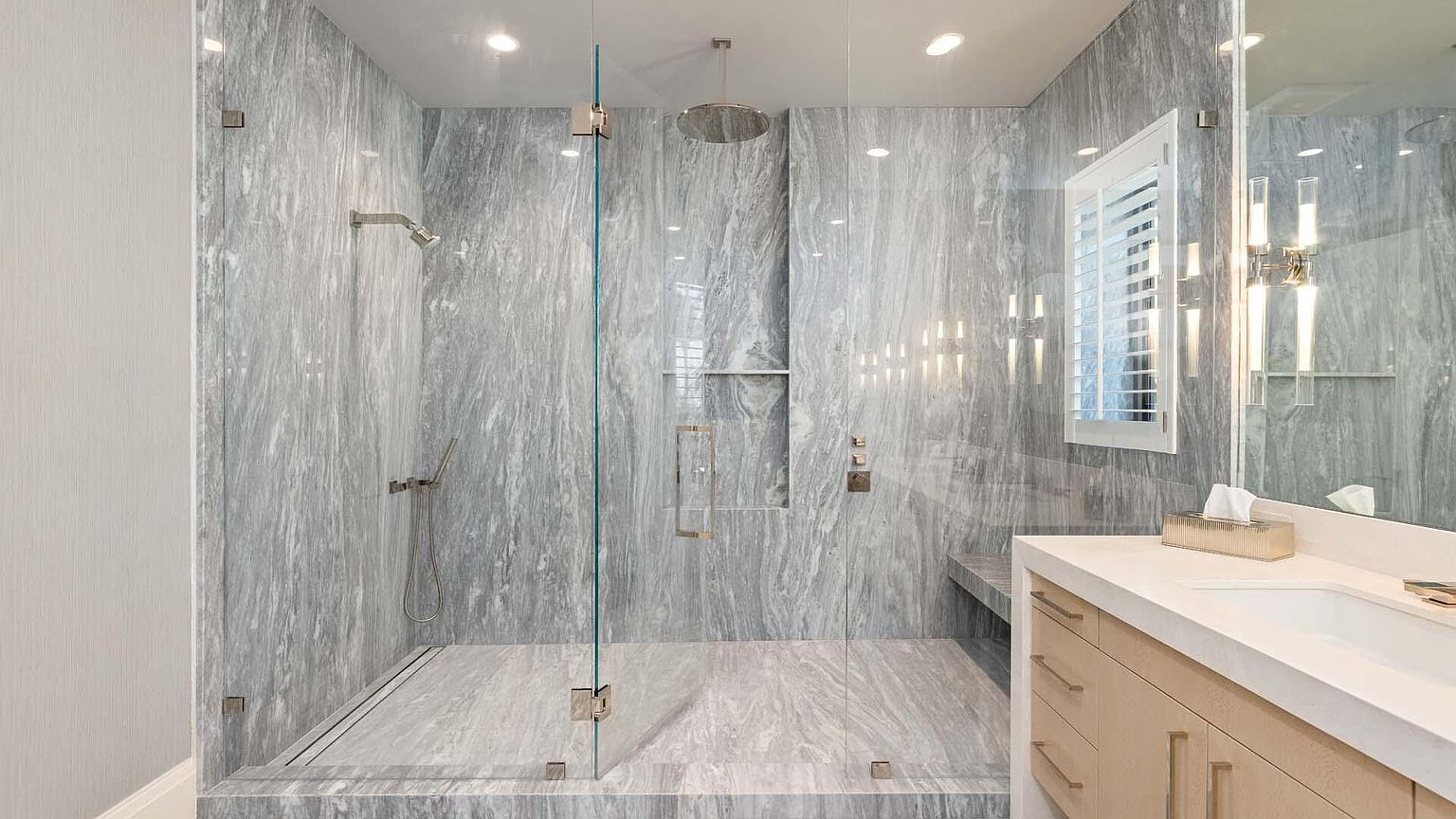 This is a luxurious primary bathroom featuring a spacious walk-in shower enclosed with glass panels, showcasing elegant gray marble walls and flooring. The shower is equipped with a rainfall showerhead and a handheld sprayer. A modern vanity with light wood cabinetry and a white countertop complements the space, while a window with shutters provides natural light.
