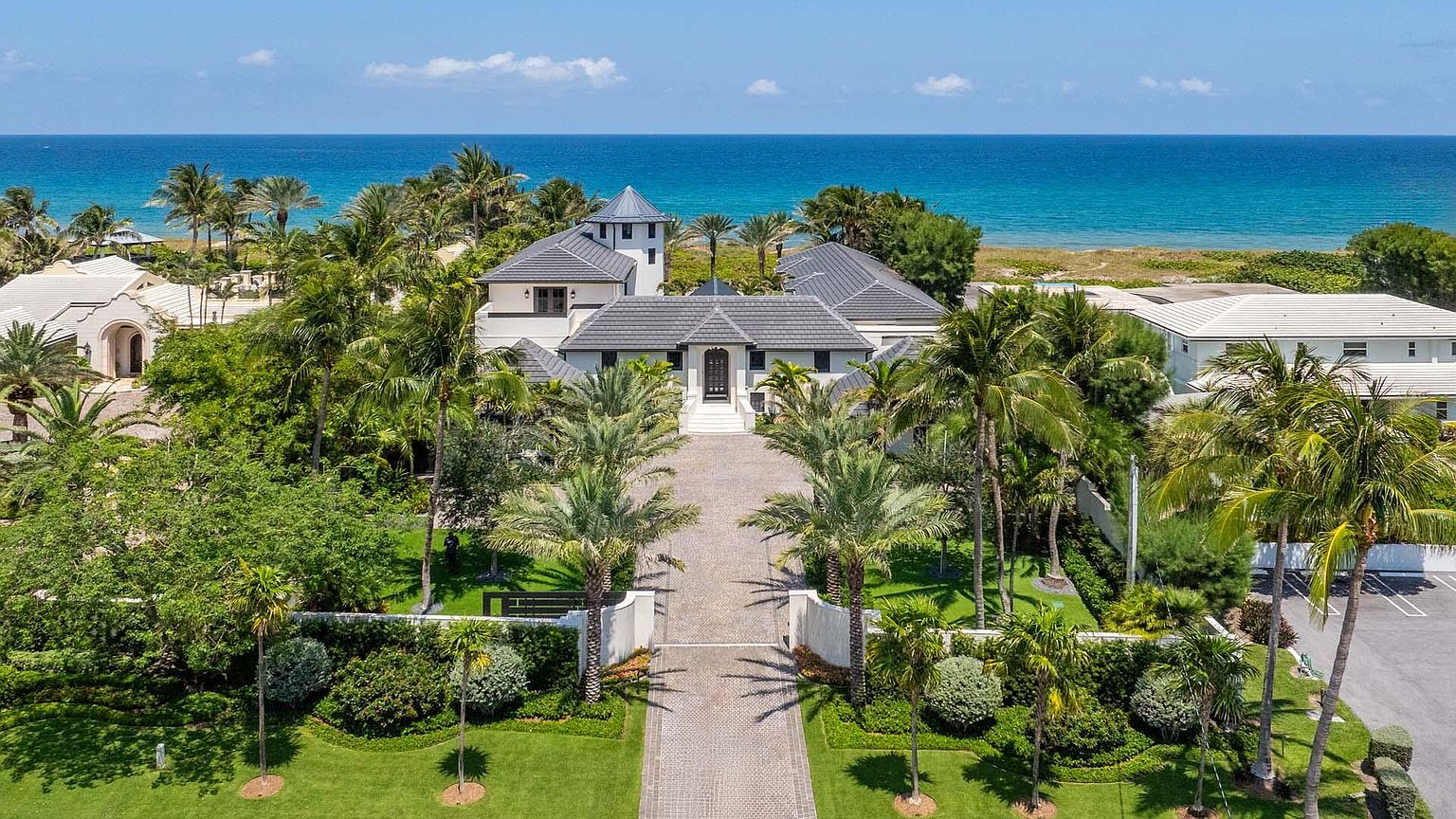 This aerial view showcases a luxurious beachfront property with a grand entrance, meticulously manicured lawns, and mature palm trees. The house features a modern architectural style with a gray tiled roof and a prominent tower element. The turquoise ocean provides a stunning backdrop, emphasizing the property's prime location and exclusivity.