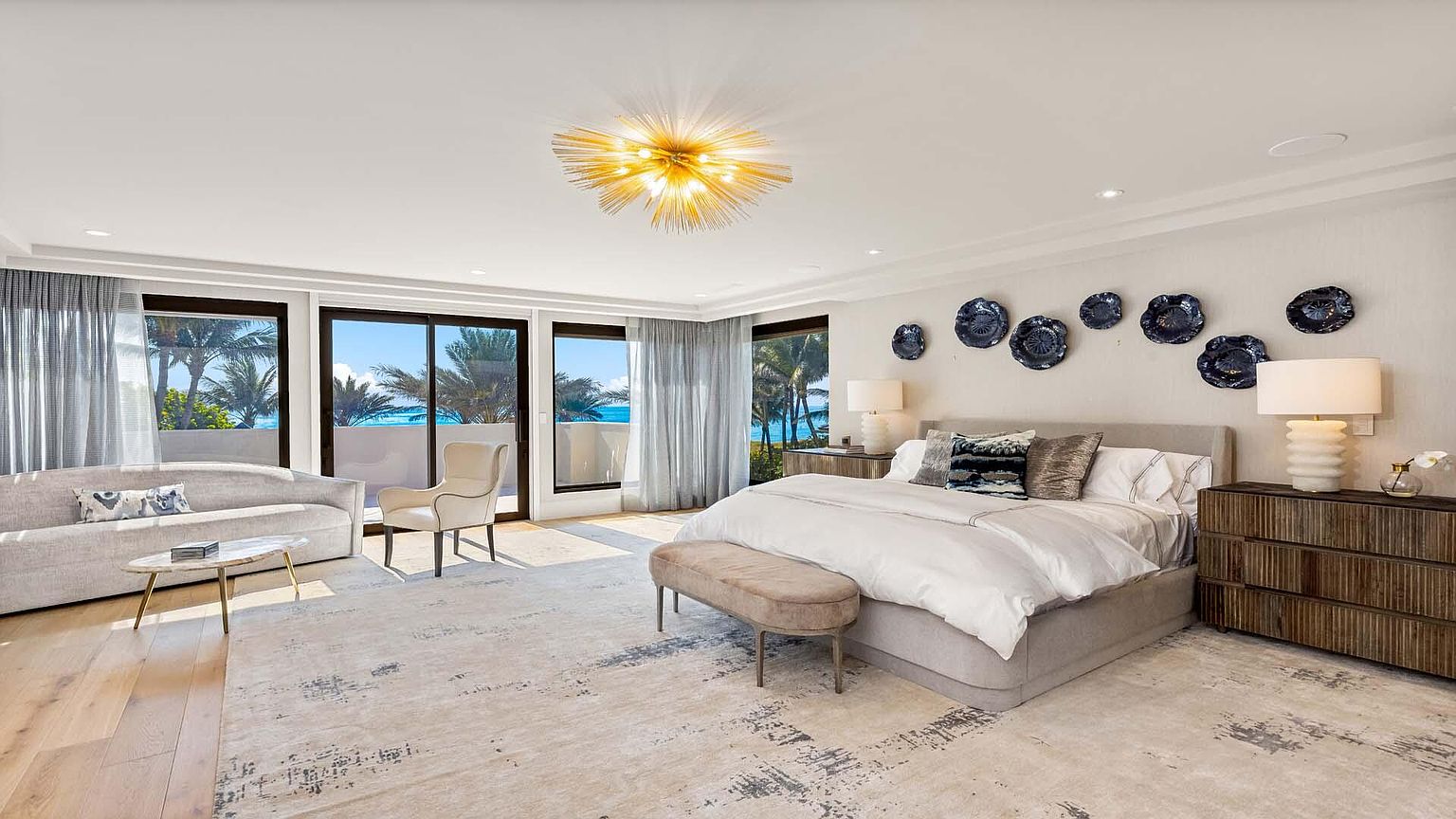 This is a bright and spacious primary bedroom featuring a large bed with neutral linens, complemented by a stylish bench and side tables. The room boasts large windows offering ocean views, a modern light fixture, and decorative wall plates, creating a luxurious and serene atmosphere. The color palette is soft and inviting, enhancing the room's overall appeal.