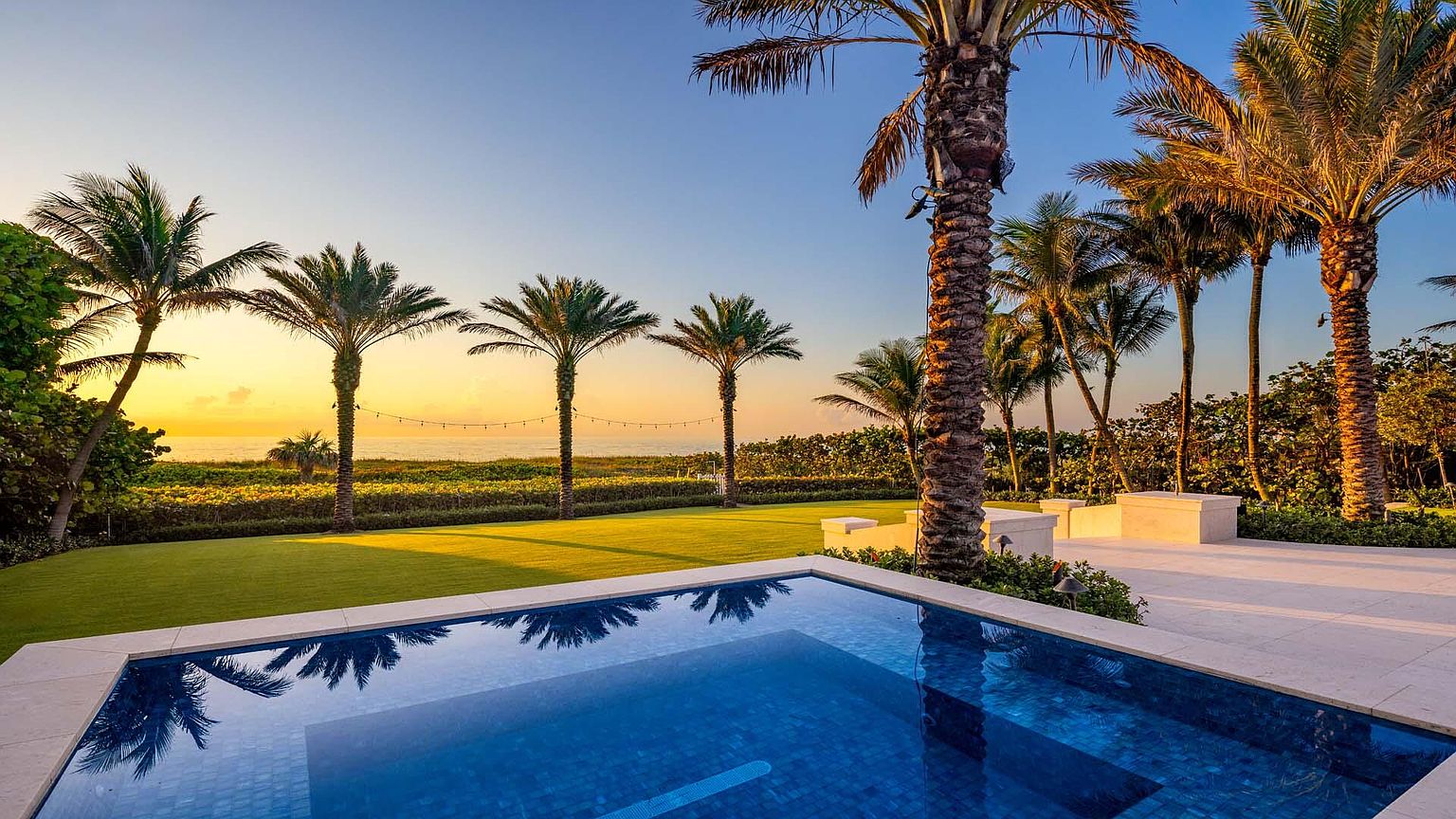 This image showcases a luxurious pool area with a stunning view of the ocean at sunset. The pool features blue mosaic tiles and a light-colored stone surround. Palm trees line the property, adding to the tropical ambiance and creating a sense of privacy and exclusivity. The perspective is from a low angle, emphasizing the pool and the expansive view beyond.