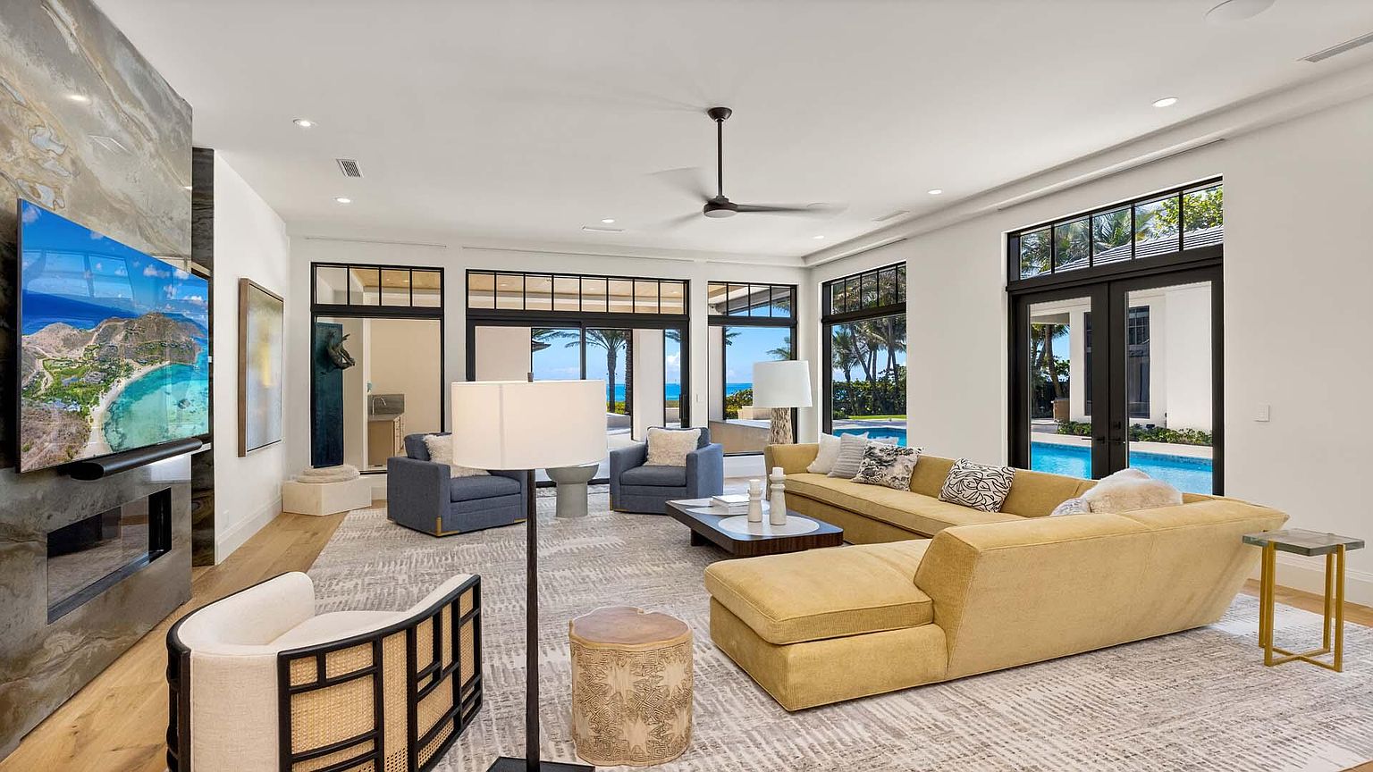 This is a bright and airy living room featuring a large yellow sectional sofa, two blue armchairs, and a patterned area rug. The room has large windows and doors that offer views of the pool and ocean. A modern fireplace with a large television above it adds a focal point to the space.