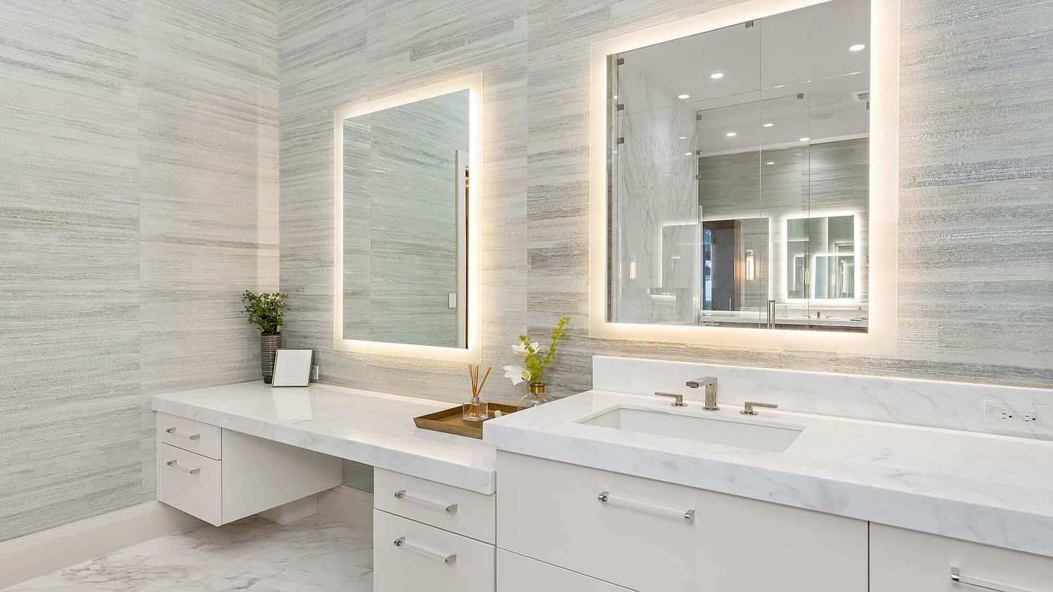 This is a well-lit primary bathroom featuring a double vanity with white marble countertops and cabinetry. Two large, illuminated mirrors hang above the vanities, reflecting the clean and modern design. The walls are covered in a light-colored, horizontally-striped tile, and the floor appears to be marble, creating a luxurious and serene atmosphere.