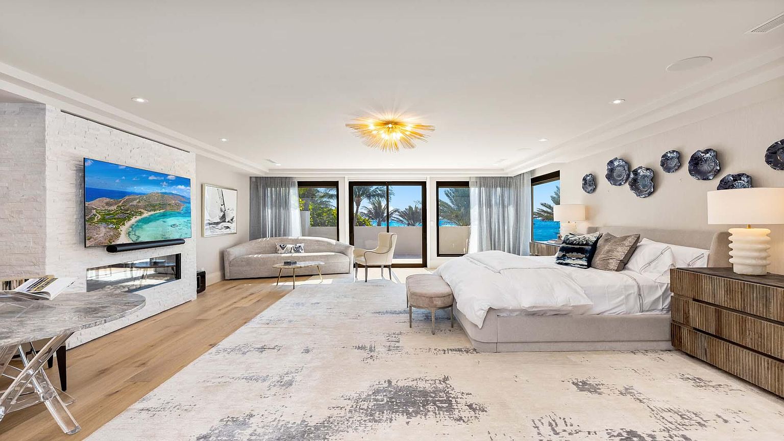 This is a spacious primary bedroom featuring a large bed with a neutral-toned headboard and white bedding. The room has a modern aesthetic with a decorative wall arrangement, a fireplace with a mounted TV, and floor-to-ceiling windows offering natural light. The flooring is light wood, and a patterned area rug adds texture to the space.
