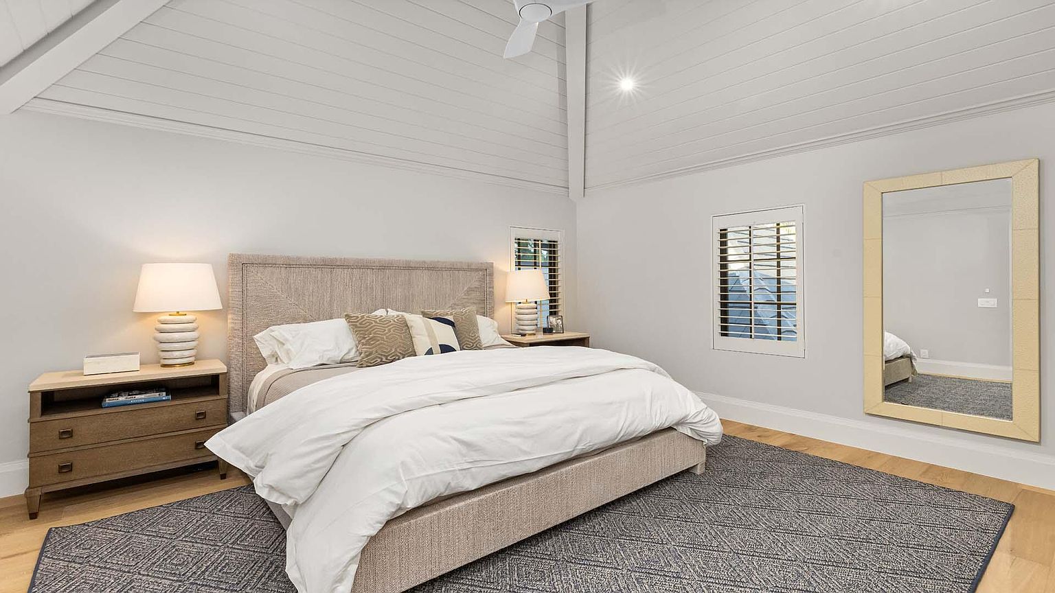 This is a bright and airy primary bedroom featuring a neutral color palette. The room includes a large bed with a woven headboard, matching nightstands with lamps, and a large mirror. The ceiling is vaulted with white wood paneling, and a patterned rug adds texture to the hardwood floor.