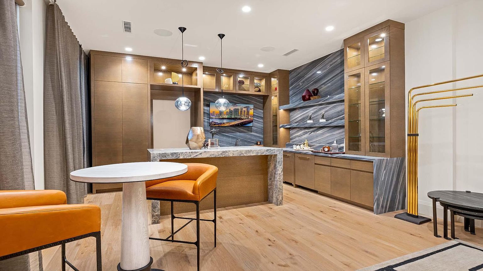 This interior shot showcases a sophisticated wine cellar or bar area, featuring custom cabinetry with glass displays, a marble countertop bar, and stylish pendant lighting. The space is designed with a modern aesthetic, incorporating a muted color palette and sleek finishes, creating an inviting and luxurious atmosphere for entertaining.