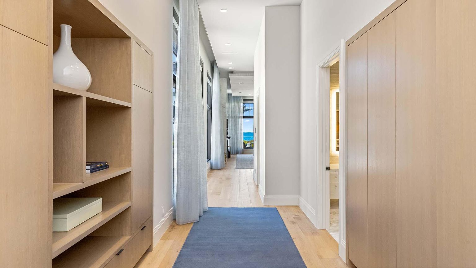 This is an interior shot of a hallway featuring light wood flooring and built-in cabinetry on both sides. A blue runner rug stretches down the center of the hallway, leading towards a window with a view of the ocean. The hallway is well-lit with recessed lighting, and a doorway to a bathroom is visible on the right.