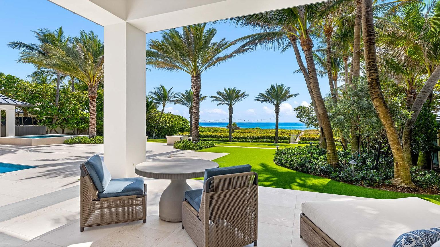 This image showcases a luxurious outdoor patio area with stylish furniture, including comfortable chairs and a modern round table. The patio overlooks a beautifully manicured lawn with palm trees and a glimpse of the ocean in the background, creating a serene and inviting atmosphere. The design emphasizes a seamless transition between indoor and outdoor living, perfect for relaxation and entertainment.