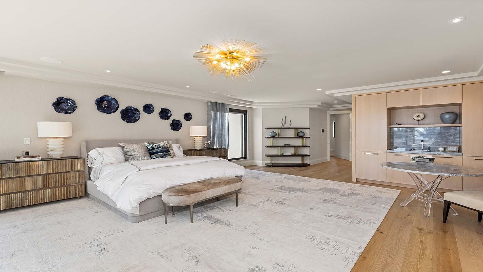 This is a spacious primary bedroom featuring a large bed with neutral-toned bedding, flanked by nightstands and lamps. The room is decorated with blue decorative wall art and a striking gold chandelier. A light-colored rug covers most of the floor, and there is a seating area with a small table and chairs, creating a luxurious and comfortable atmosphere.