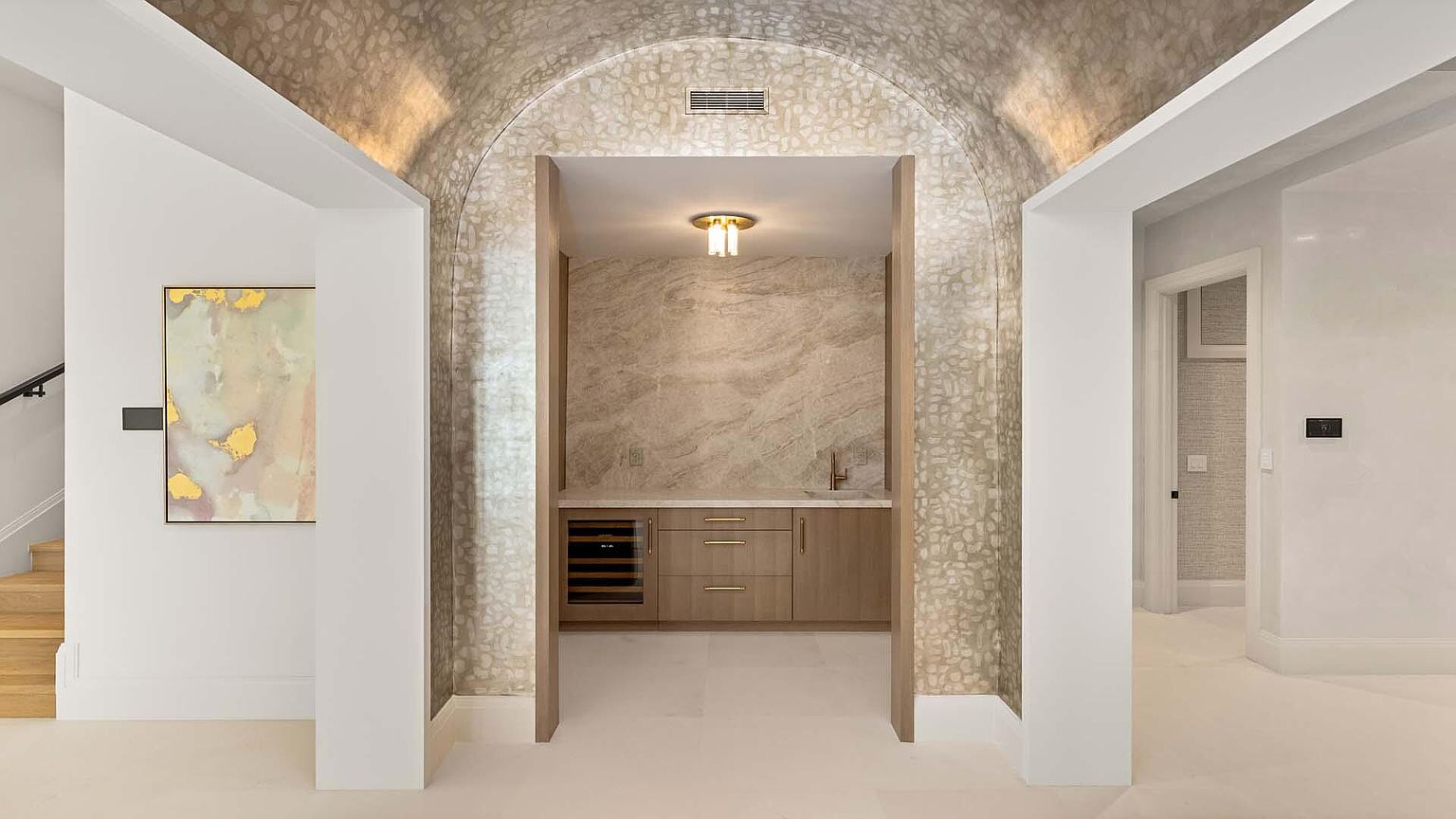 This interior shot showcases a hallway with an arched alcove leading to a bar area. The hallway features white walls and trim, with a painting on the left and a glimpse of stairs. The bar area has a marble countertop, wooden cabinets, and a wine cooler, all illuminated by a modern light fixture.
