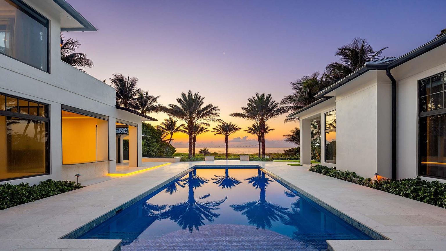 This image showcases a luxurious outdoor pool area at dusk, framed by modern white buildings on either side. Palm trees line the horizon, reflecting beautifully in the pool's still water, creating a serene and inviting atmosphere. The scene is bathed in the warm hues of the sunset, enhancing the property's appeal as a tranquil retreat.