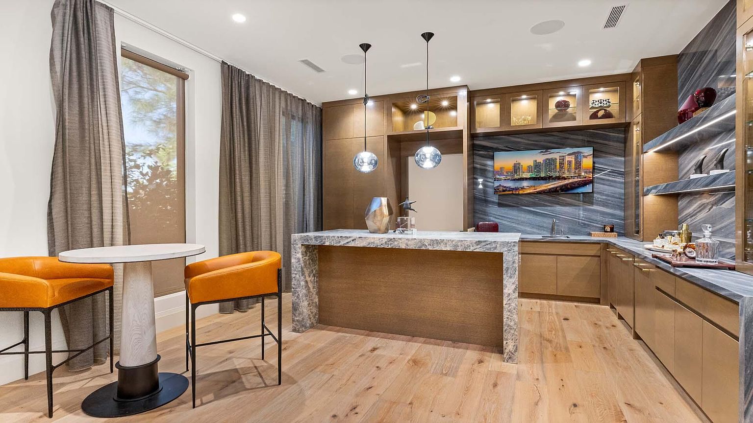 This is an interior shot of a modern wine cellar or bar area. It features a marble-topped bar with wood paneling, complemented by orange bar stools and a small table. The space includes built-in cabinetry with glass displays, a television, and stylish pendant lighting, creating a sophisticated and inviting atmosphere.