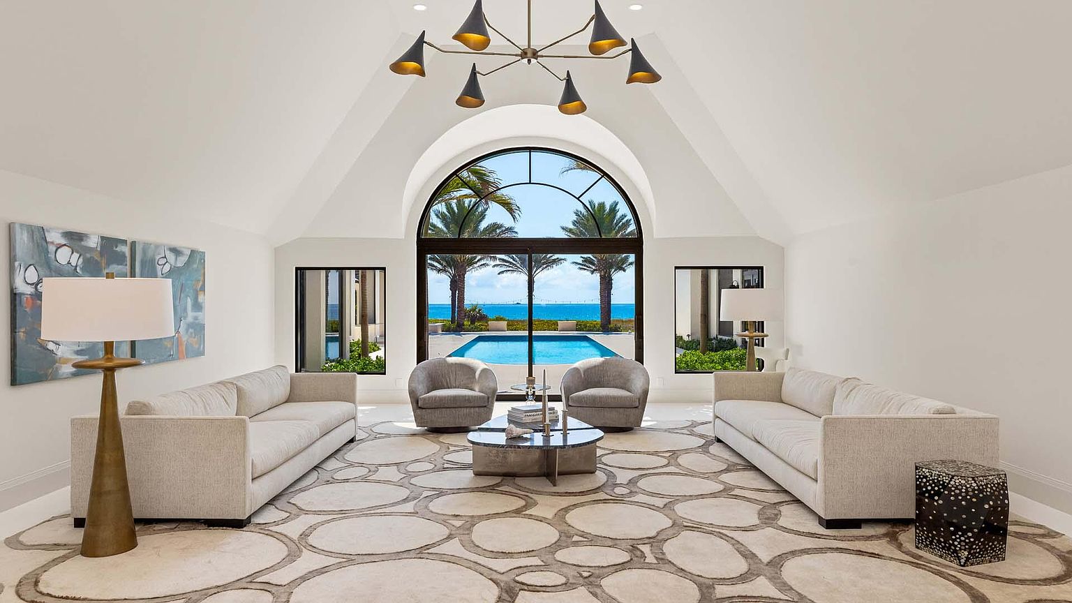 This is an interior shot of a luxurious living room featuring two light-colored sofas, two armchairs, and a unique circular patterned rug. A large arched window provides a stunning view of a pool and the ocean beyond, framed by palm trees. The room is well-lit with a modern chandelier and lamps, creating an elegant and inviting atmosphere.