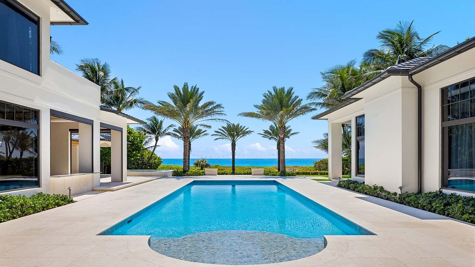 This image showcases a luxurious backyard featuring a rectangular swimming pool with a mosaic tile detail at the entry. The pool is flanked by well-manicured landscaping and palm trees, with a glimpse of the ocean in the background. The architecture of the adjacent buildings is modern and elegant, creating a serene and upscale outdoor living space.