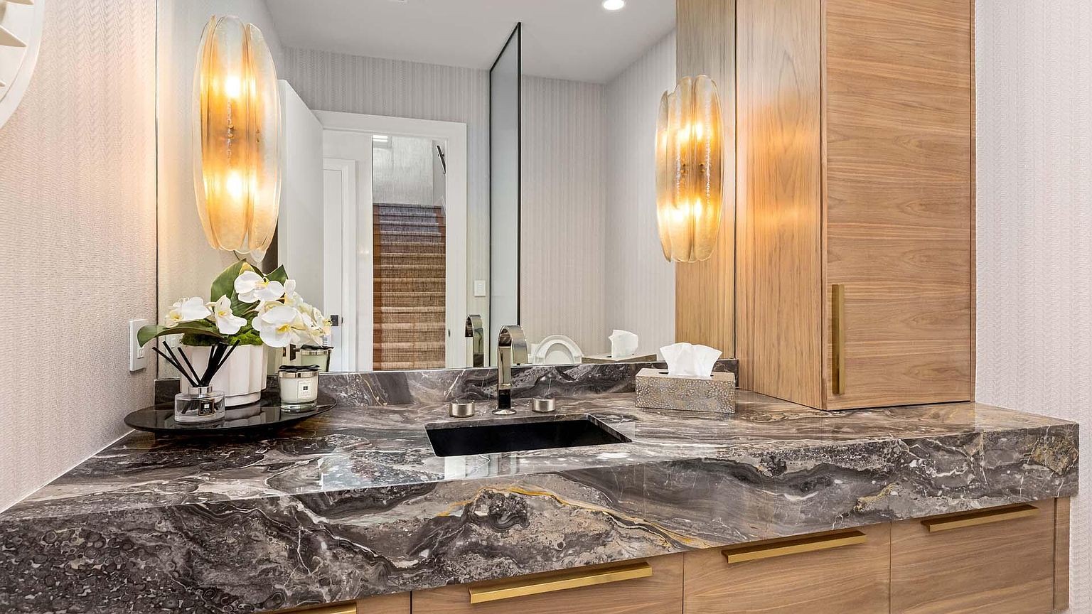 This is a stylish guest bathroom featuring a dark marble countertop with a black rectangular sink. The vanity is made of light wood with gold hardware, and the walls are covered in a subtle textured wallpaper. Two sconces provide warm lighting, and a mirror reflects the doorway leading to another room, creating a sense of depth.