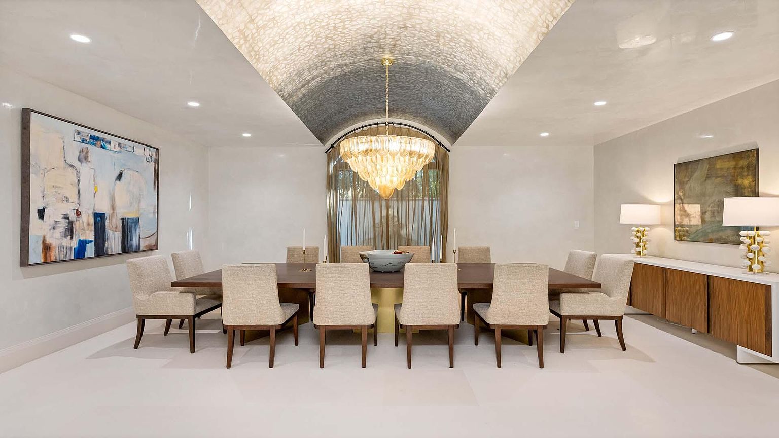 This is an elegant dining room featuring a long wooden table surrounded by upholstered chairs. A large, ornate chandelier hangs above the table, illuminating the space. Artwork adorns the walls, and a sideboard with lamps adds to the room's sophisticated ambiance. The room has a vaulted ceiling with a unique textured finish.