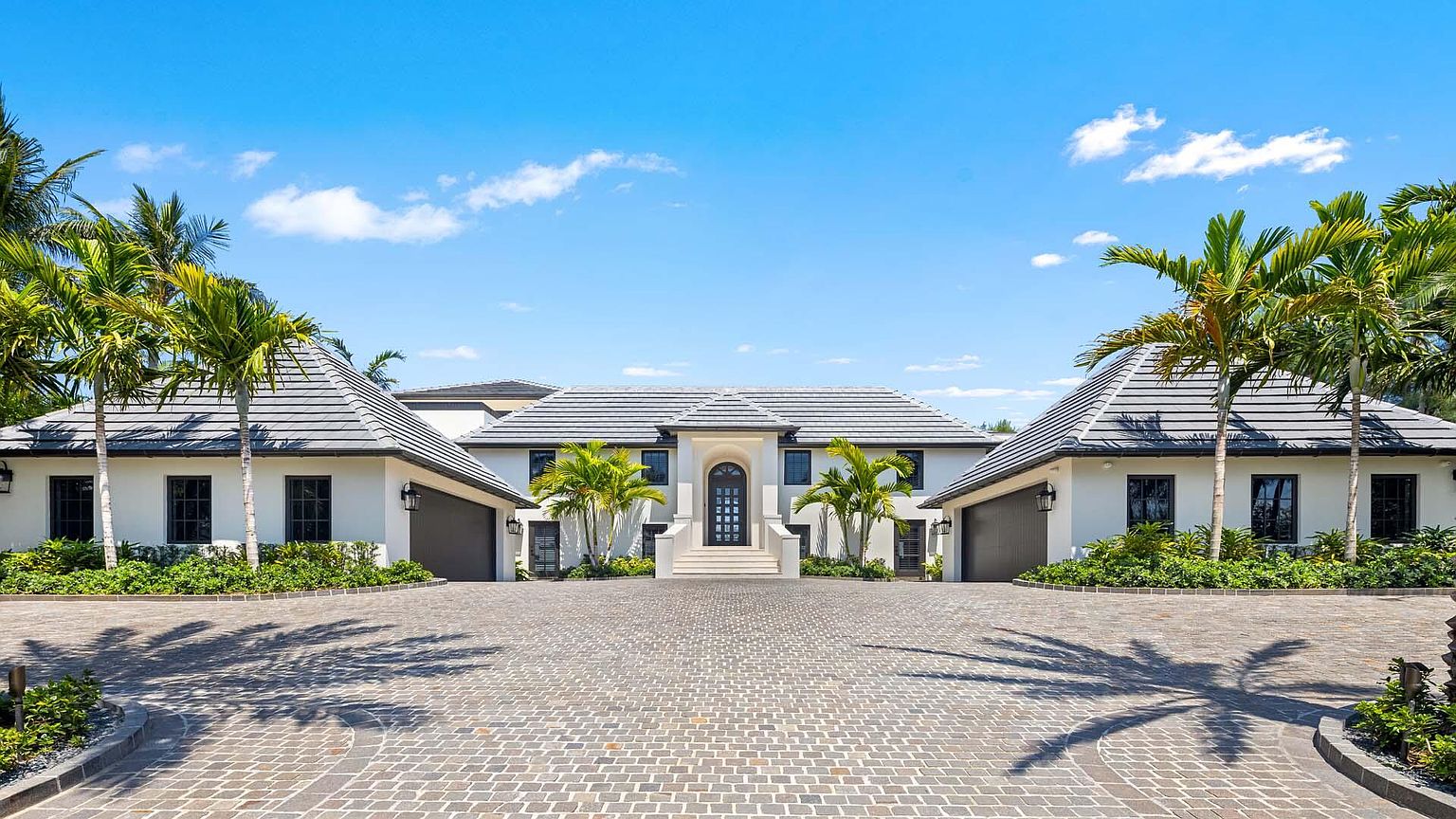 This is a symmetrical front view of a luxurious, modern home. The house features a light-colored exterior, dark-framed windows, and a gray tiled roof. Palm trees line the property, and a paved driveway leads to the entrance, creating a grand and inviting approach.