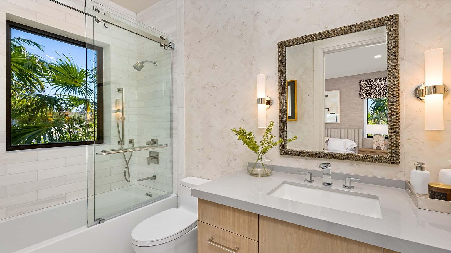 This is a well-lit bathroom featuring a modern design. The bathroom includes a bathtub with a glass shower door, a toilet, and a vanity with a light wood cabinet and a light-colored countertop. A decorative mirror hangs above the sink, and the walls are covered in a light-colored patterned tile, creating a clean and inviting space.