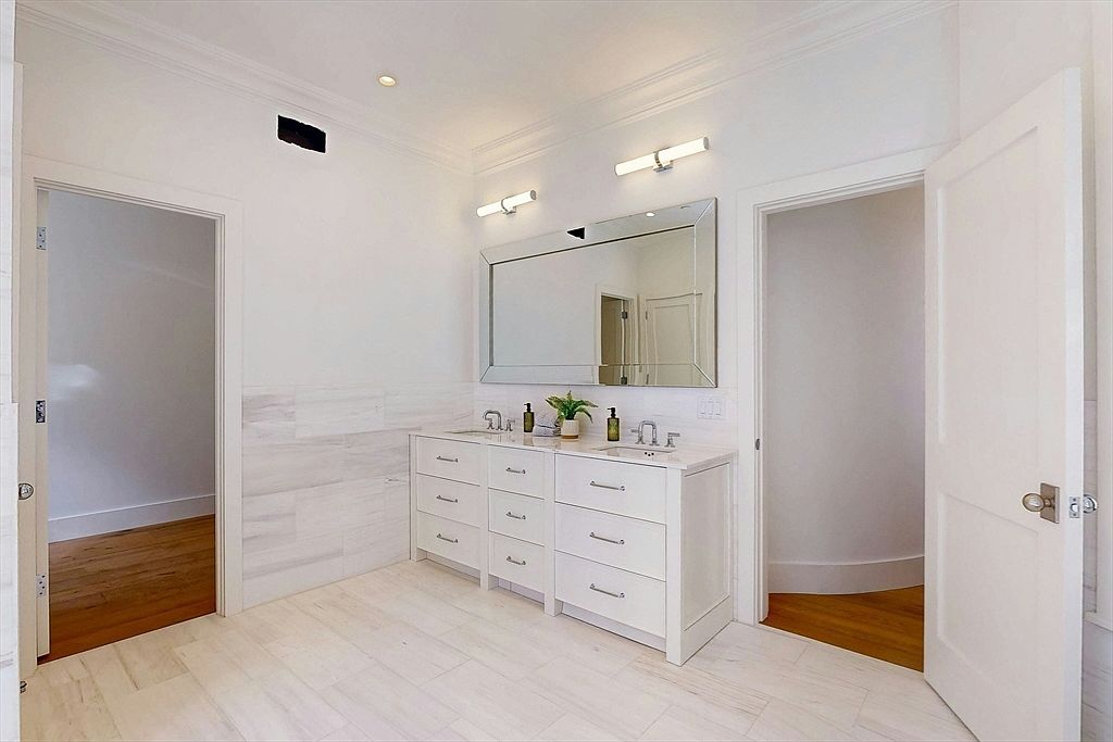 This bright and airy bathroom features a double vanity with white cabinetry and a light-toned stone countertop. A large, rectangular mirror is centered above the vanity, flanked by two modern wall-mounted light fixtures. The space is finished with light-colored tile flooring and wainscoting, creating a clean, minimalist aesthetic with doorways leading to adjacent rooms.