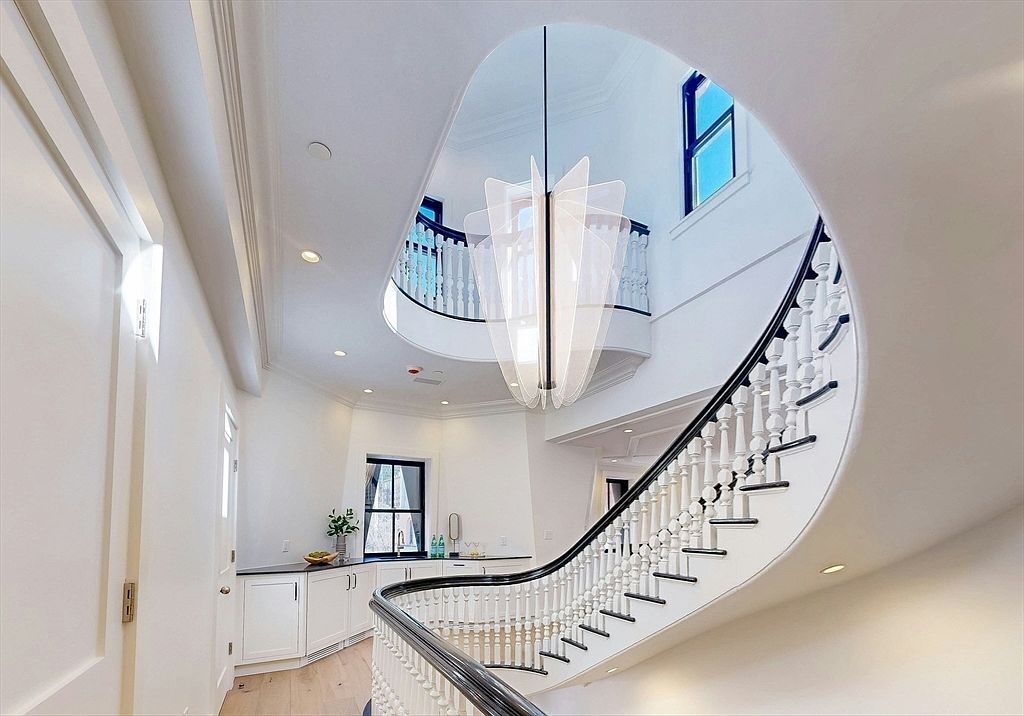 This elegant foyer features a dramatic curved staircase with a dark wood handrail and white balusters, leading to an upper landing. A striking, modern sculptural chandelier hangs from the ceiling, serving as a focal point within the open, light-filled space. In the background, a small wet bar area with white cabinetry and a dark countertop is visible, adding a touch of convenience to the sophisticated entryway.