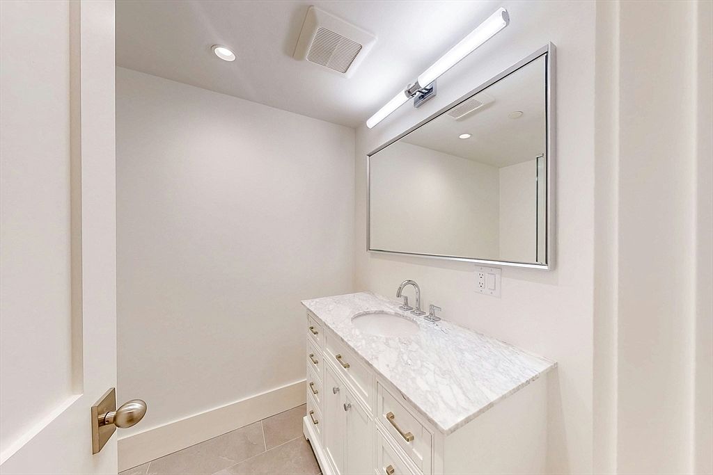 This clean and bright powder room features a white vanity topped with a light-toned stone counter and an undermount sink. A large, rectangular mirror with a metallic frame hangs above the vanity, illuminated by a sleek, modern light fixture. The space is finished with neutral-toned floor tiles and crisp white walls, creating a minimalist and polished aesthetic.
