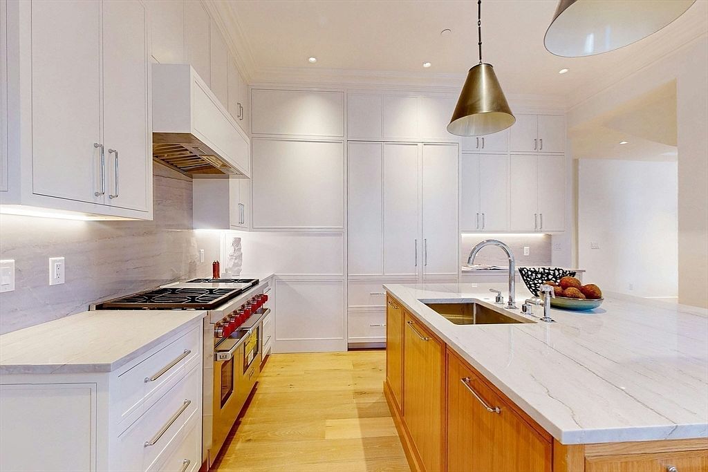 This bright, modern kitchen features a clean aesthetic with white cabinetry contrasted by a warm-toned wood kitchen island. The space is equipped with a professional-grade range, a stone-topped island with an integrated sink, and a large metallic pendant light hanging above. The perspective is a wide-angle shot from the kitchen entrance, showcasing the seamless flow between the cooking area and the central island.