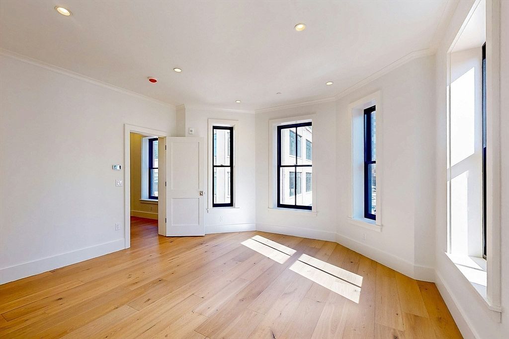 This bright and airy room features light wood flooring and crisp white walls, creating a clean, minimalist aesthetic. Three large windows with dark frames allow ample natural light to fill the space, while a doorway leads into an adjacent room. The room's neutral palette and simple architectural lines offer a versatile canvas for various interior design styles.