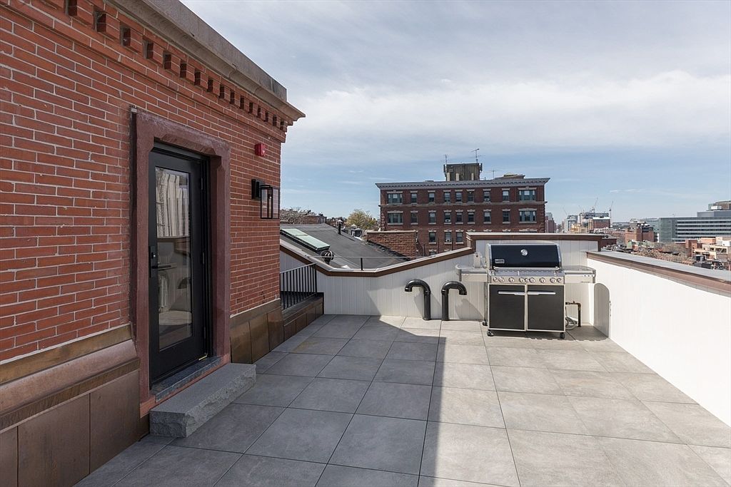 This rooftop deck offers a spacious outdoor living area paved with large, neutral-toned tiles. The space features a brick wall on one side, a black-framed glass door leading inside, and a stainless steel grill positioned against a white railing. The perspective provides an elevated view of the surrounding urban cityscape under a bright, partly cloudy sky.
