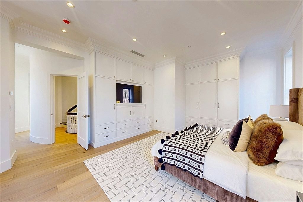 This bright and spacious primary bedroom features light wood flooring and a large, patterned area rug centered beneath the bed. A wall of custom white cabinetry provides extensive storage and integrates a television, while a doorway leads to a curved hallway with a view of a staircase. The room is decorated with a plush bed adorned with textured pillows and a patterned throw, creating a clean and sophisticated atmosphere.