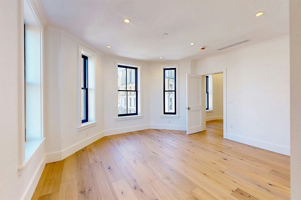This bright, minimalist living space features light wood flooring and crisp white walls, creating an airy and open atmosphere. The room is defined by a curved wall section with multiple tall windows that allow for abundant natural light and views of the surrounding neighborhood. A white door leads into an adjacent room, emphasizing the clean, modern architectural lines of the home.