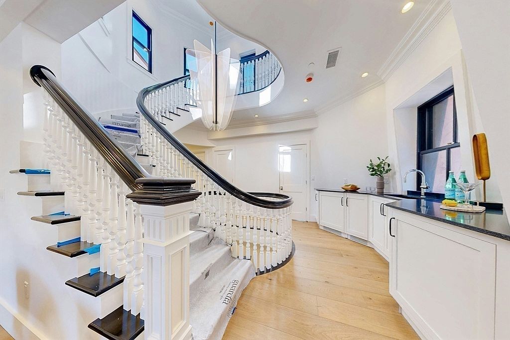 This elegant entryway features a grand, curved staircase with a dark wood handrail and white balusters, serving as the focal point of the space. To the right, a sleek wet bar area is integrated into the hallway, showcasing white cabinetry, a dark stone countertop, and a small sink. The light wood flooring and bright, neutral walls create an airy, sophisticated atmosphere, while a modern, sculptural chandelier hangs from the ceiling to add a touch of contemporary flair.