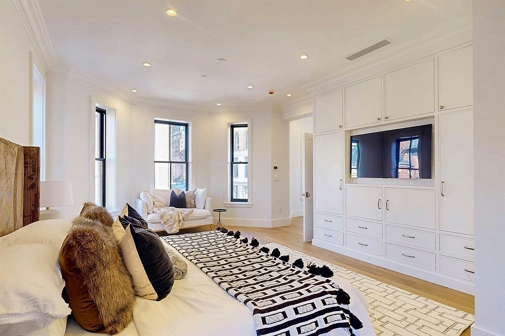 This bright and airy primary bedroom features a large bed with textured pillows and a patterned throw blanket, positioned in front of a wall of custom white built-in cabinetry that integrates a television. The room is illuminated by recessed lighting and natural light streaming through three tall windows, which frame a cozy seating area with a white loveseat. The space is finished with light wood flooring, creating a clean, sophisticated, and inviting atmosphere.