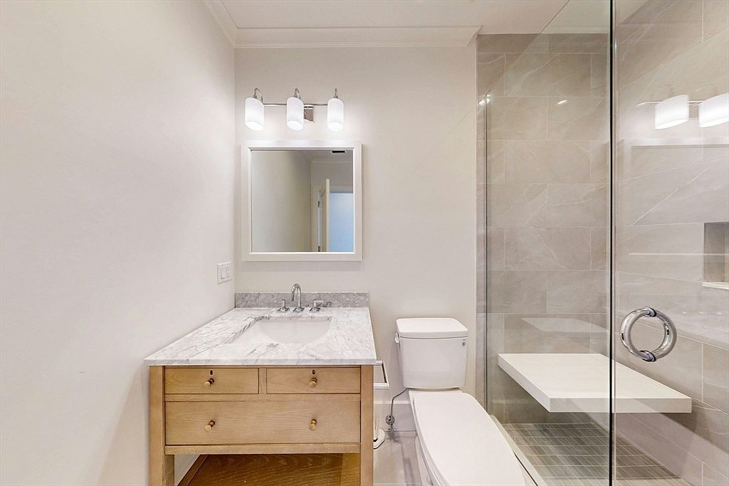 This clean and modern bathroom features a light wood vanity topped with a stone counter and an undermount sink. To the right, a glass-enclosed shower area includes a built-in stone bench and neutral-toned wall tiles. The space is illuminated by a three-light fixture above a rectangular mirror, creating a bright and functional atmosphere.
