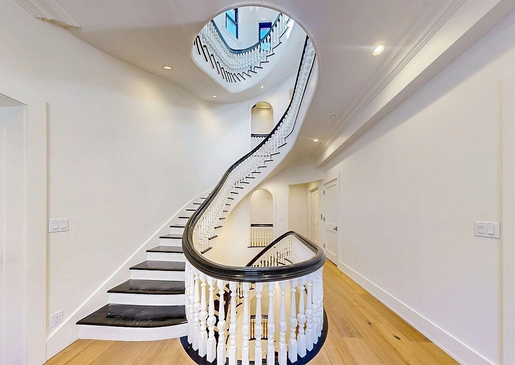 This grand foyer features a striking, curved staircase with dark-stained treads and white balusters that winds upward through an open, multi-level atrium. The space is finished with crisp white walls and light-toned wood flooring, creating a bright and airy atmosphere. The perspective from the base of the stairs emphasizes the architectural elegance and the vertical flow of the home's design.