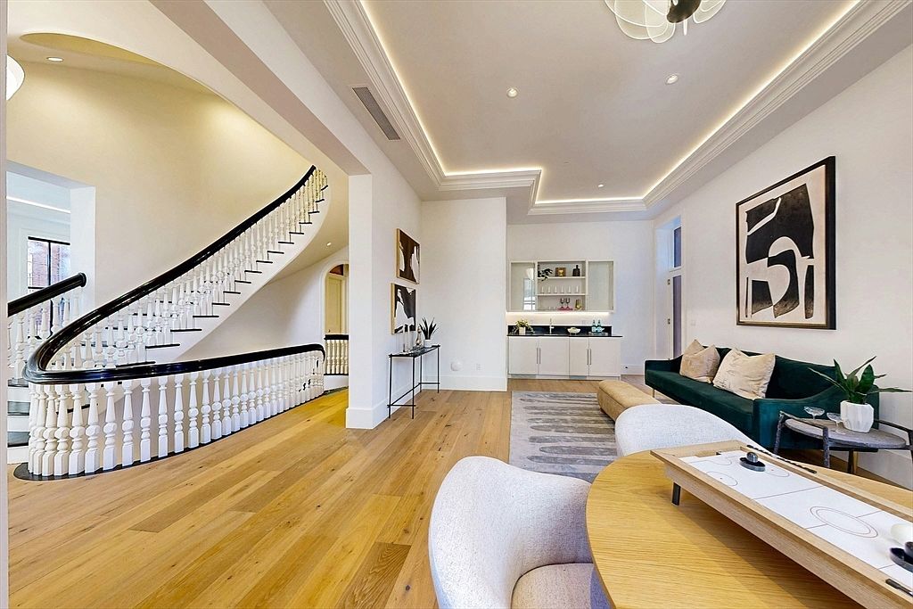 This elegant open-concept living space features light wood flooring and a dramatic curved staircase with a dark handrail. The room is anchored by a deep green sofa and a round wooden table, complemented by a wet bar area with built-in cabinetry in the background. The space is illuminated by recessed lighting and a modern chandelier, creating a sophisticated and airy atmosphere.
